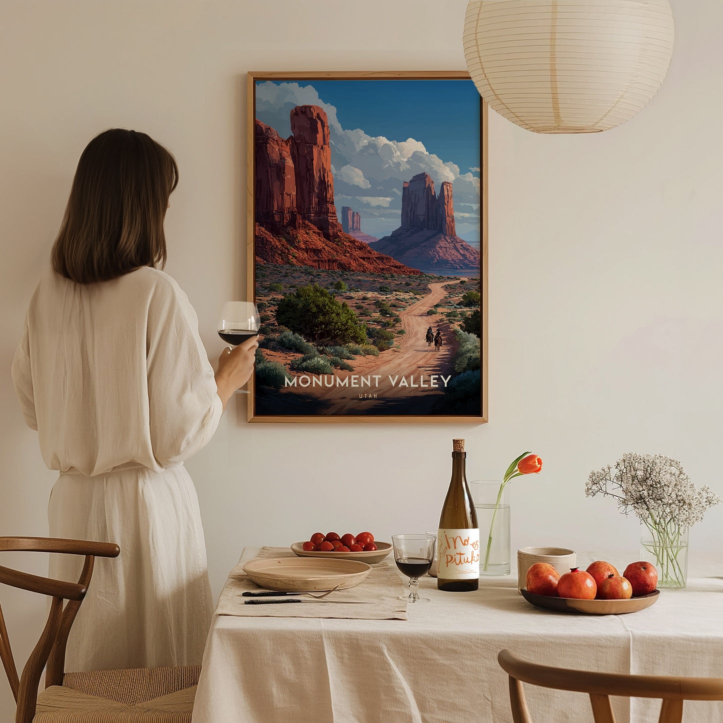 A woman in a white dress holds a glass of wine, admiring a Monument Valley poster on the wall. The table is set with fruit, wine, and flowers, creating a.