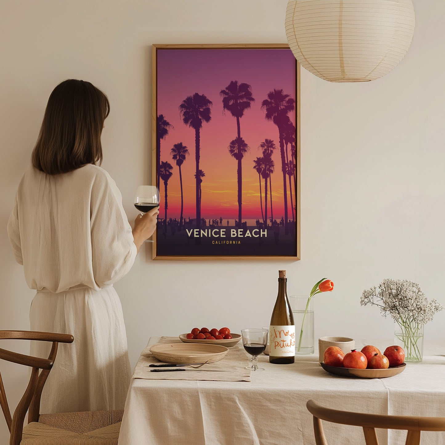 A woman in a white dress admires a framed Venice Beach sunset poster with palm trees. A table with wine, fruit, and flowers is set in the foreground.