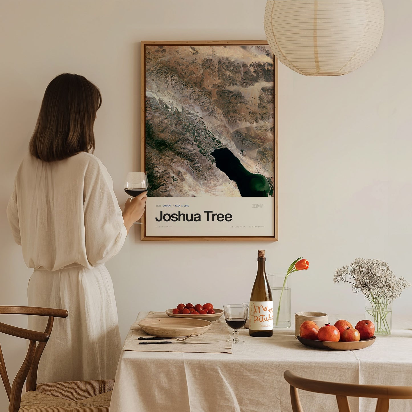 A woman in a light-colored outfit stands holding a glass of wine, gazing at a framed poster titled Joshua Tree.