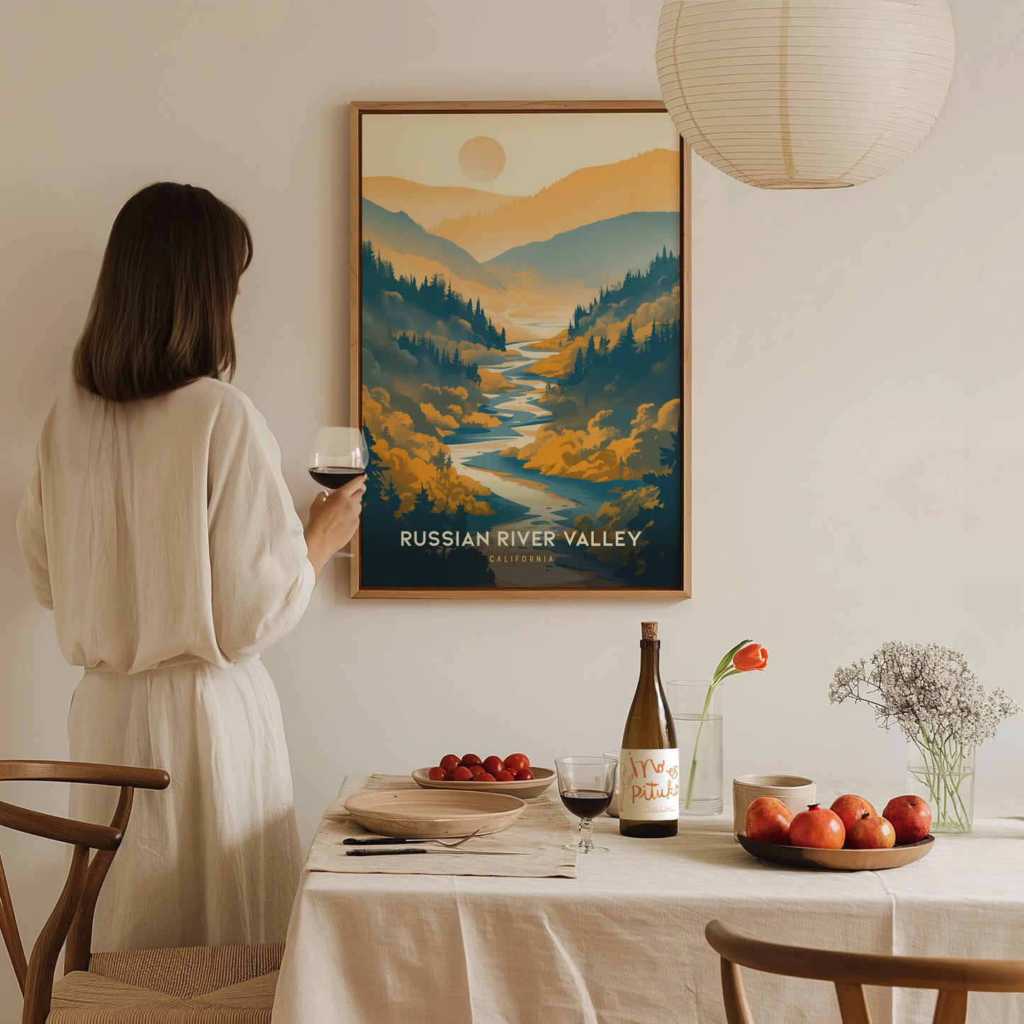 A woman in a white dress admires a Russian River Valley poster while holding a glass of wine. A table is set with fruits, wine, and a flower vase.