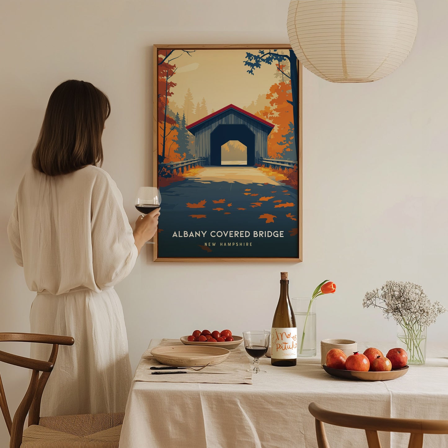 A woman stands in a cozy dining area, holding a glass of wine and admiring a framed poster of the Albany Covered Bridge in New Hampshire.