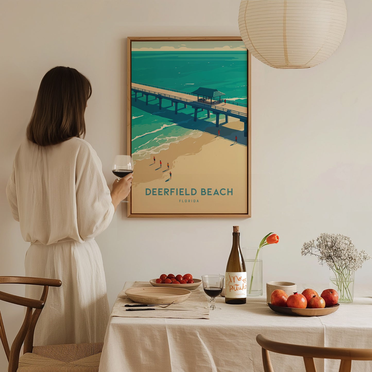 A woman in a white dress admires a Deerfield Beach poster on the wall. A dining table with wine, fruit, and flowers is set in the foreground.