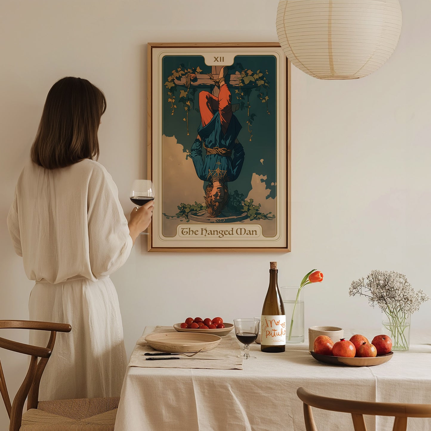 A woman holding a glass of wine gazes at a framed Hanged Man Tarot poster on the wall. A table set with fruit, flowers, and a wine bottle is in.