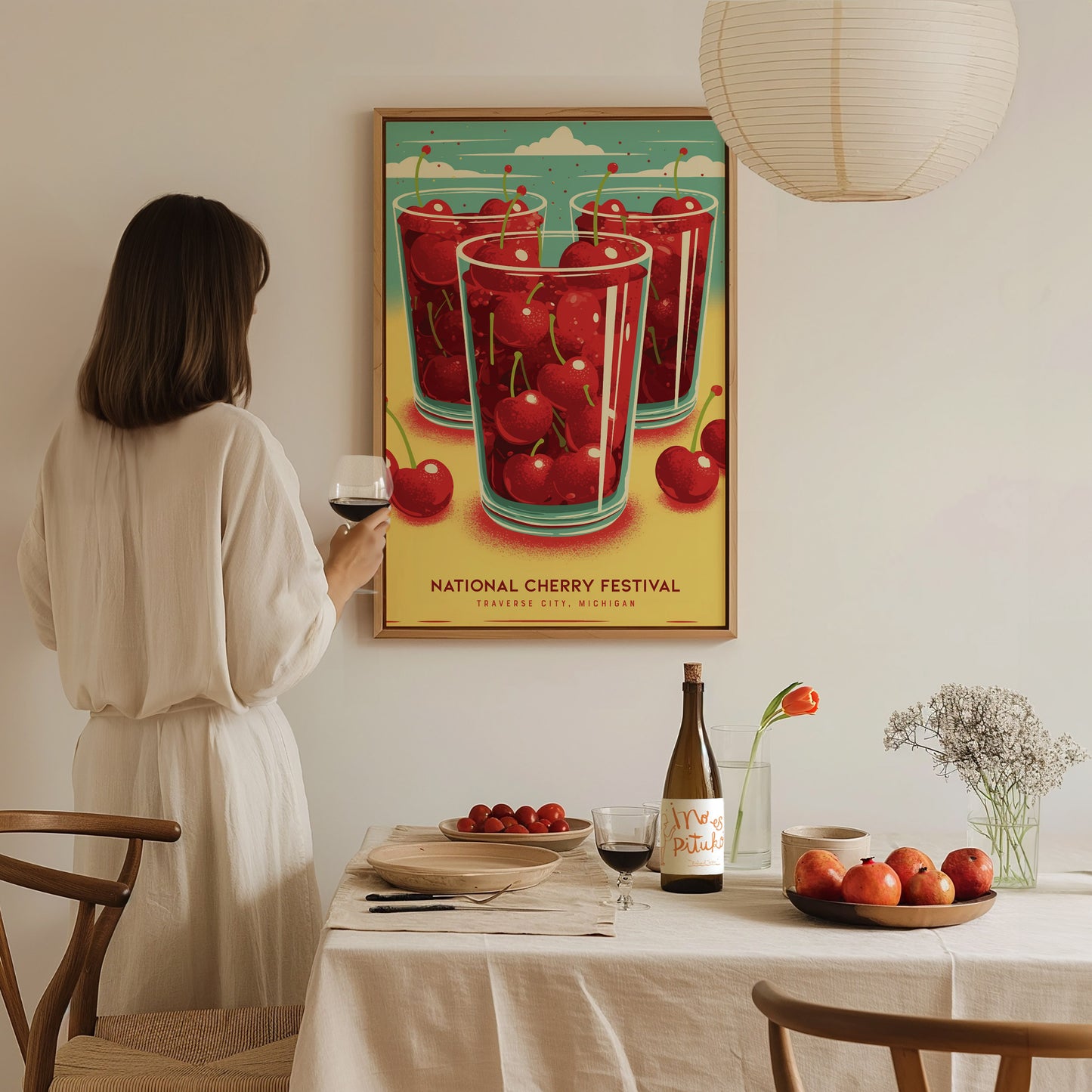 A woman in a white dress admires a colorful National Cherry Festival poster featuring cherries. The table is set with wine, fresh cherries, and a vase with flowers.