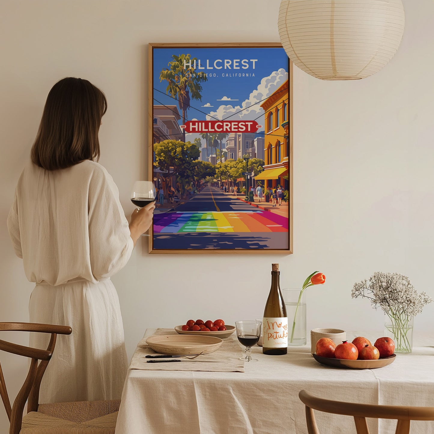 A person in a white dress holds a wine glass while admiring a colorful Hillcrest poster on the wall. A table with fruit, wine, and flowers is in the foreground.