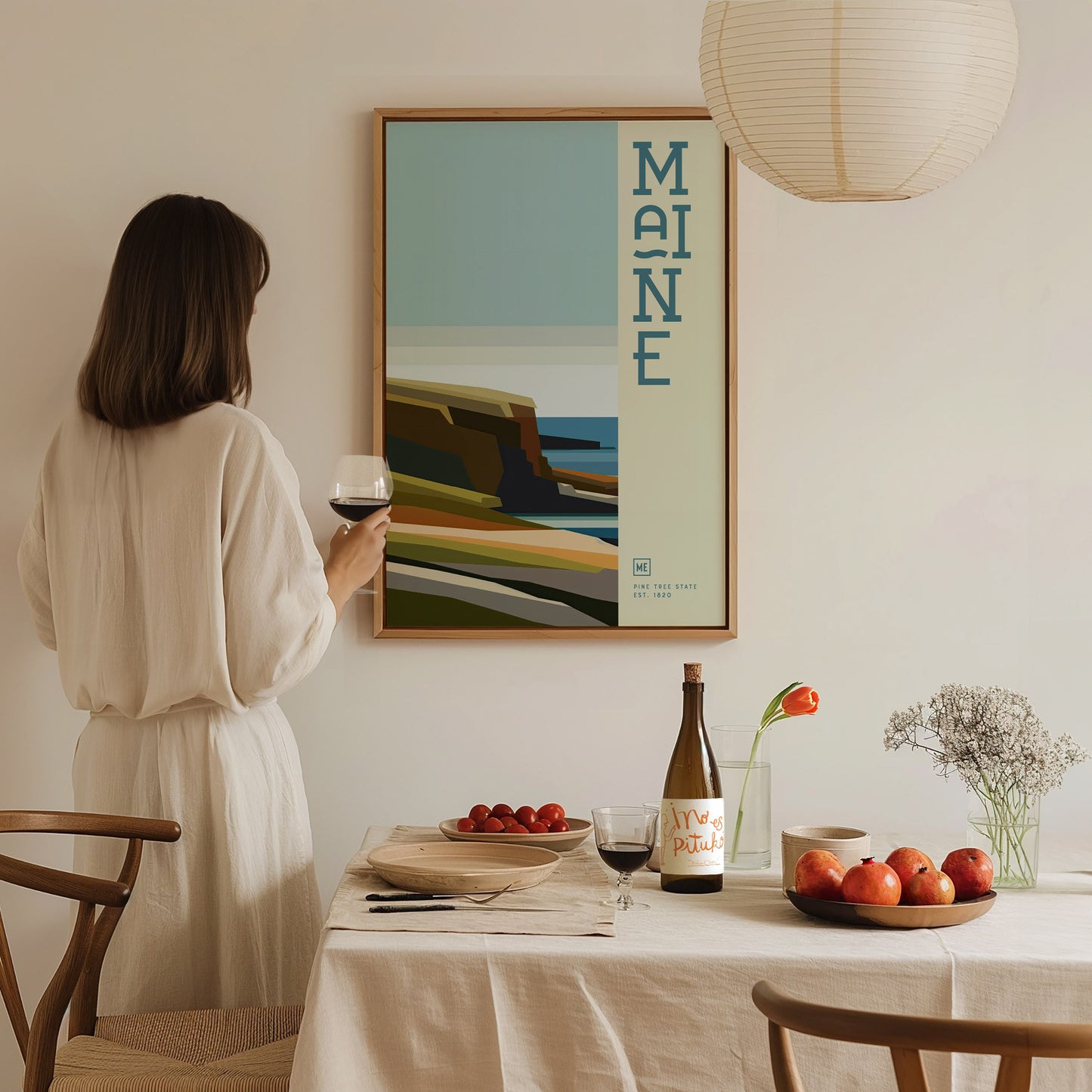 A woman in a beige dress holds a glass of wine while admiring a colorful Maine poster on the wall. A dining table with fruits and wine is in the.