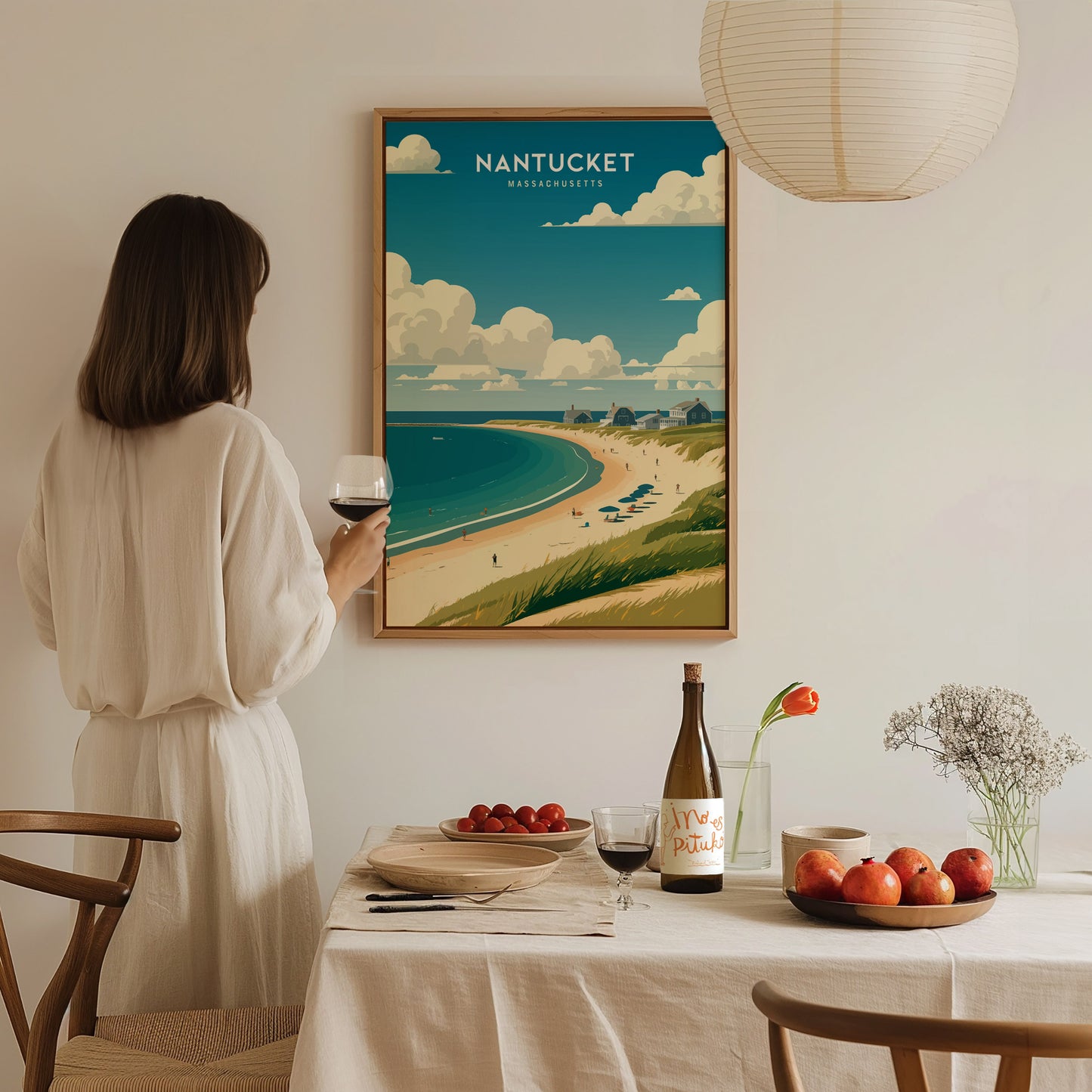 A woman in a white dress holds a wine glass, admiring a framed Nantucket poster on a wall above a dining table with fruit and wine.