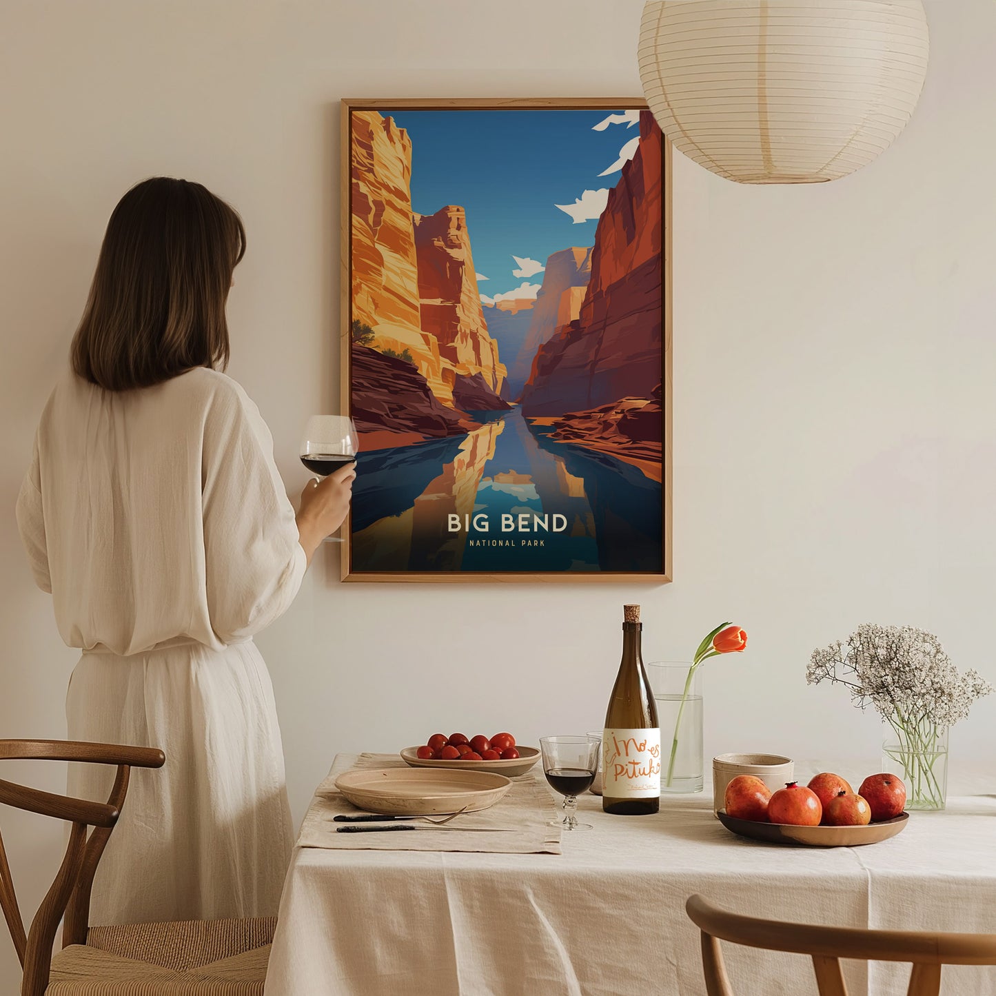 A woman in a white dress admires a framed Big Bend National Park poster on a wall. A table set with wine, fruit, and flowers complements the serene atmosphere.