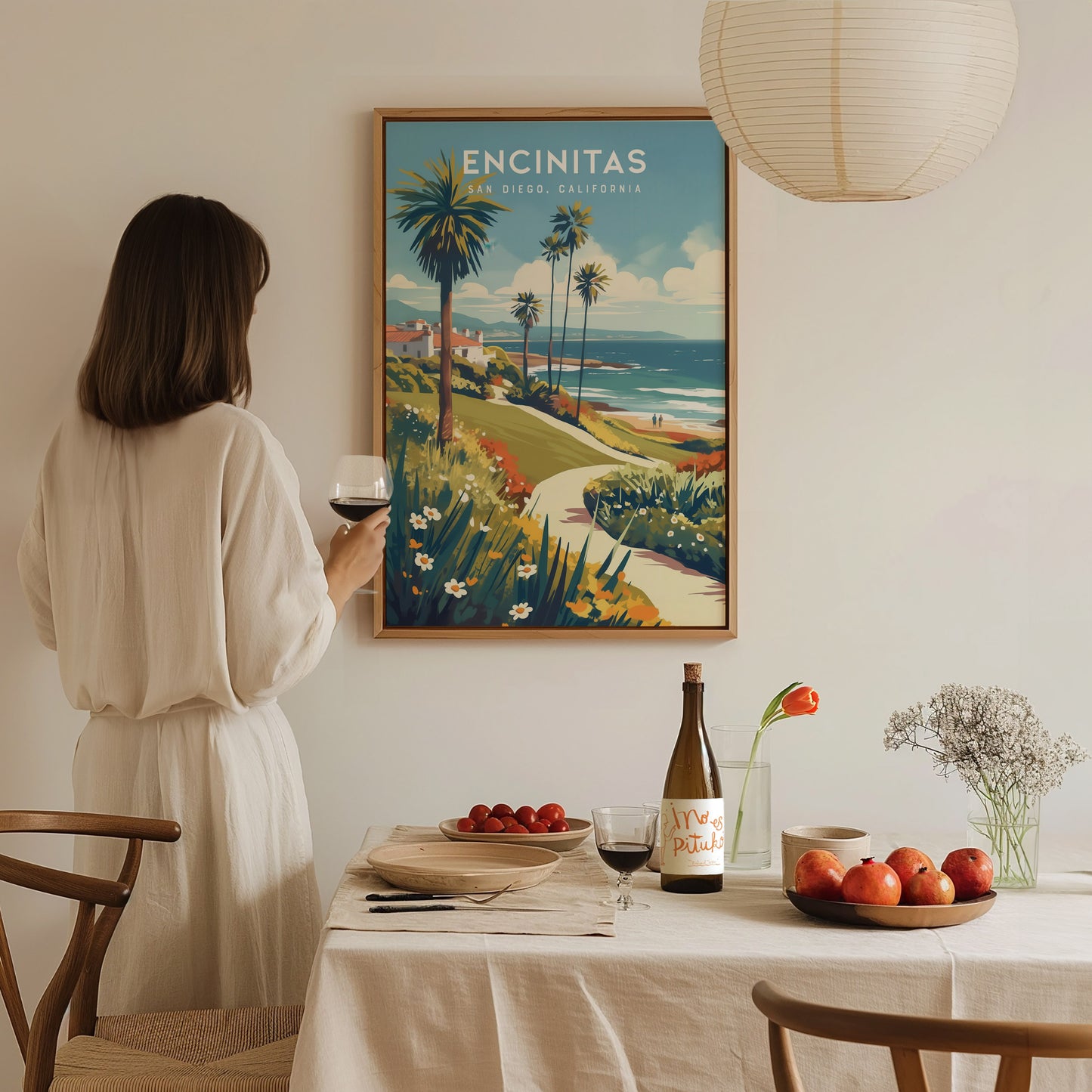 A woman in a white dress holds a glass of wine, admiring a framed Encinitas, California poster on the wall. A dining table with fruit and wine is in the.