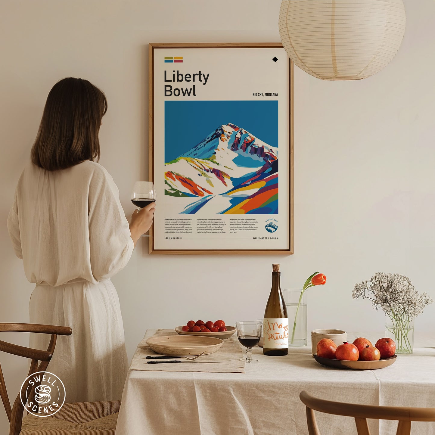 A woman in a white dress holds a glass of wine, admiring a colorful Liberty Bowl Big Sky poster on the wall.