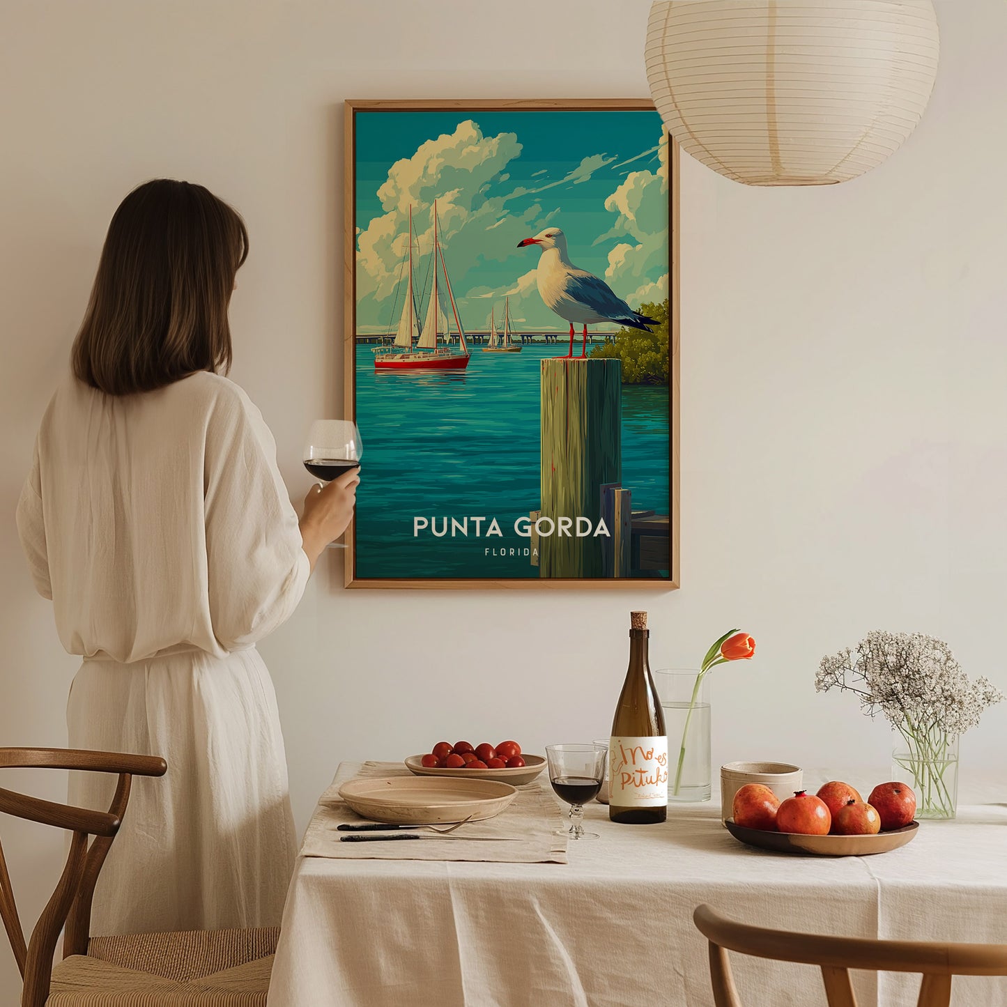 A woman in a white outfit stands beside a table set with a bottle of wine, glasses, and fruit, admiring a framed poster of Punta Gorda, Florida, featuring boats and a seagull against a vibrant sky.