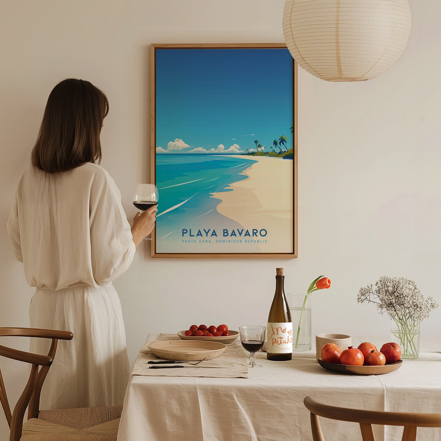 A woman in a white outfit stands near a dining table, holding a glass of wine, while admiring a framed poster of Playa Bavaro. The table is set with a bottle of wine, a plate of fruits, and decorative elements in a cozy, well-lit room.