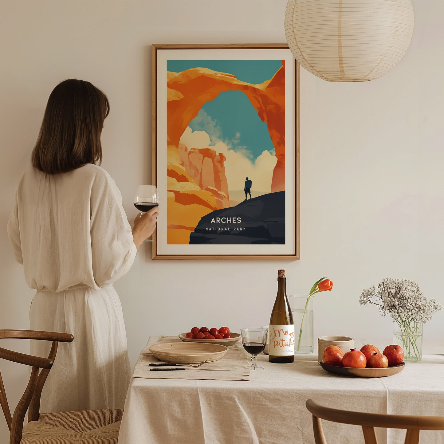 A woman in a white dress holds a wine glass while admiring a framed Arches National Park poster. A table with fruit, a bottle, and flowers is in the foreground.