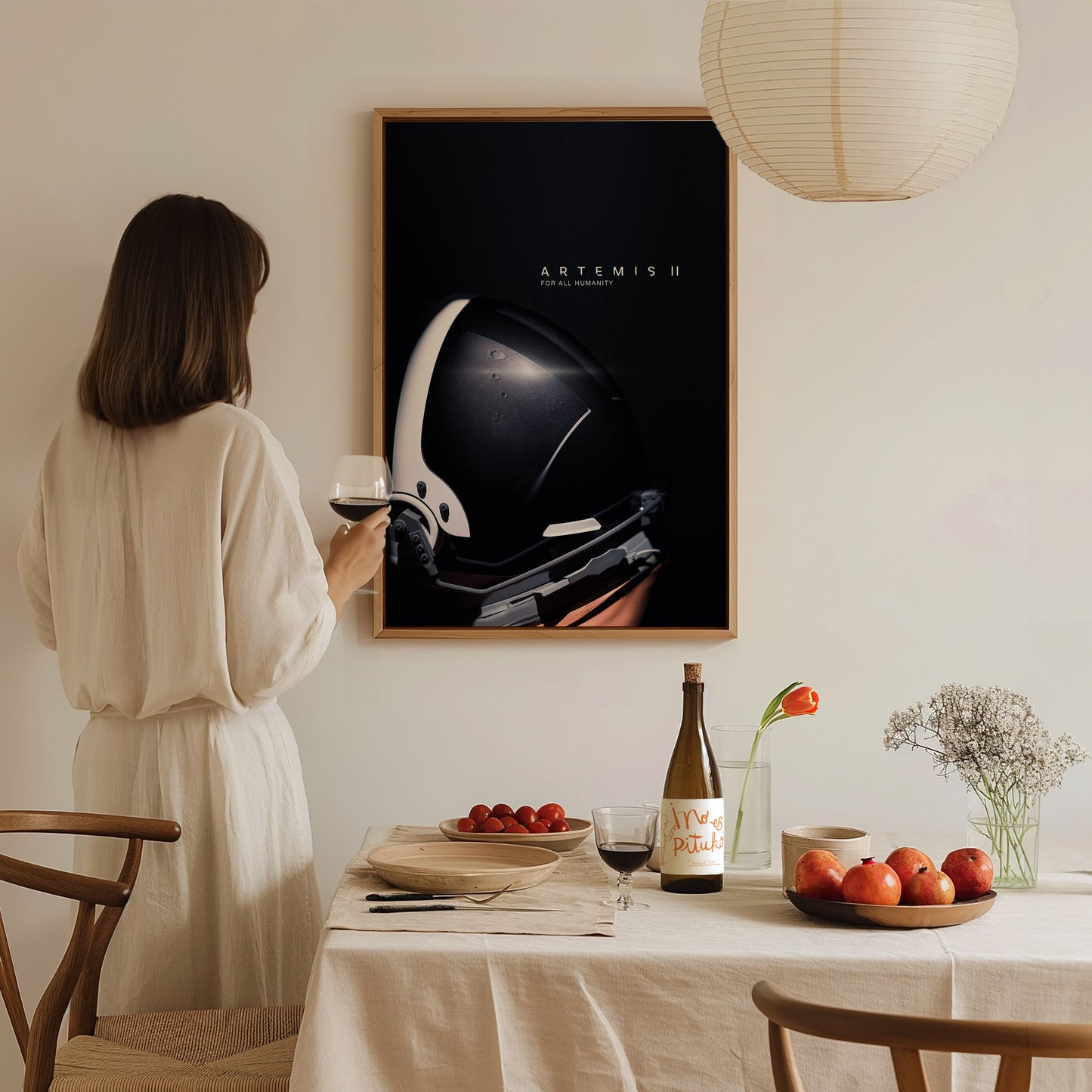 A woman in a white dress stands by a dining table, holding a glass of wine and admiring a framed poster featuring the Artemis II space helmet design. The table is set with apples, a bottle, and a vase of flowers, creating a cozy atmosphere.
