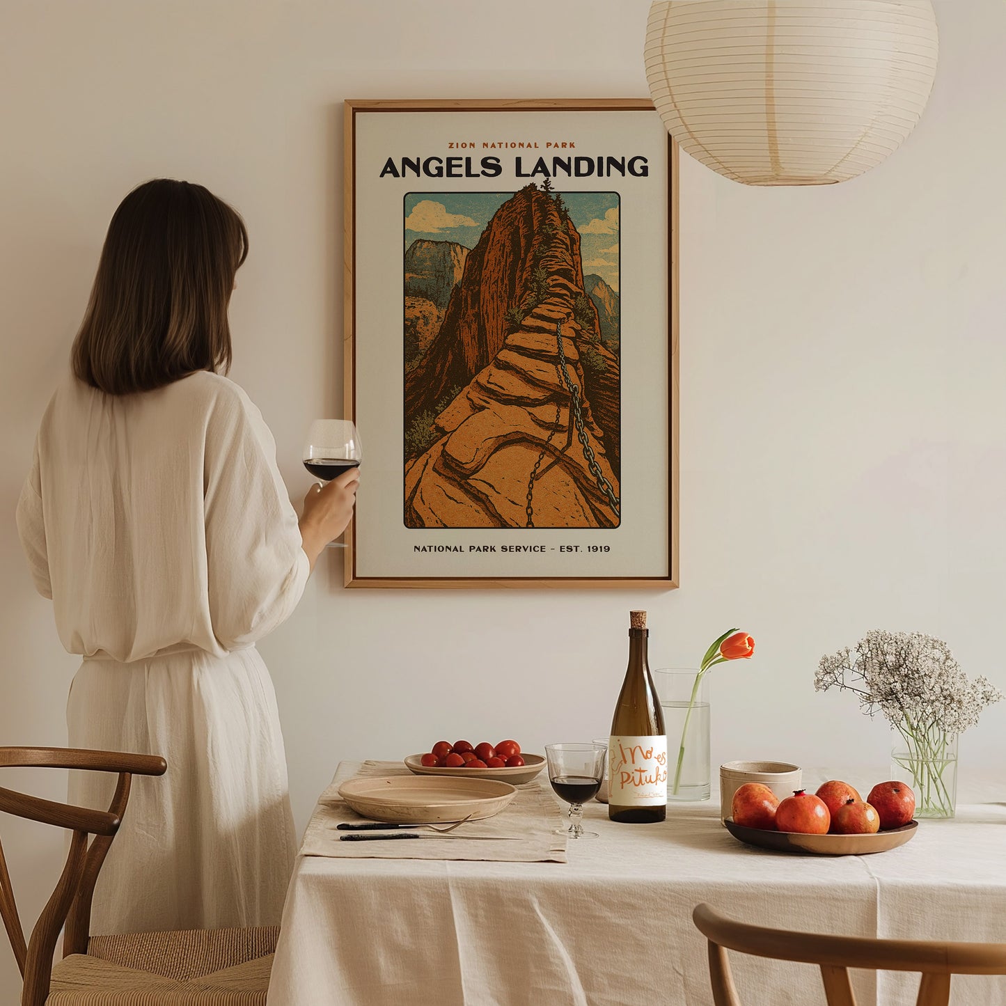 A woman in a white dress holds a glass of wine, admiring an Angels Landing poster. The table is set with fruit, a wine bottle, and a vase with flowers.