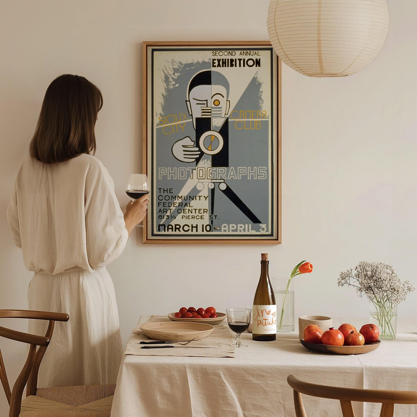 Woman in a white dress standing at a dining table with a vintage-style poster on the wall.