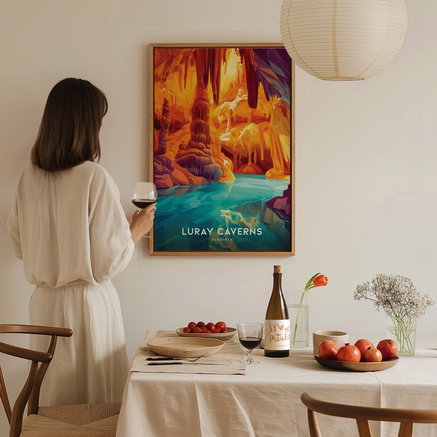 A woman in a white dress holds a glass of wine, admiring a colorful Luray Caverns poster on the wall. A table with fruit and wine is in the foreground.