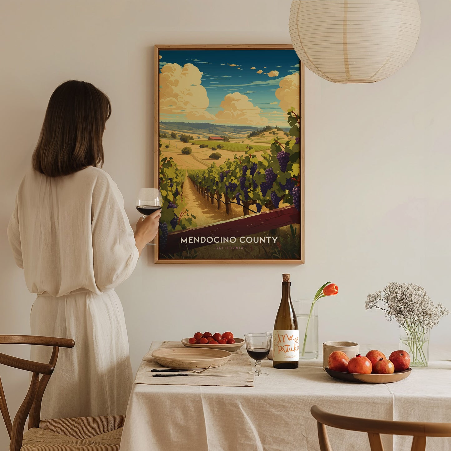 A woman in a white dress holds a glass of wine, admiring a Mendocino County poster. A table with fruits, wine, and flowers is set in front.