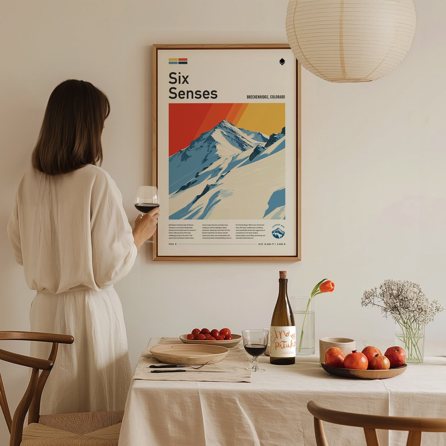 A woman in a white dress admires a framed Six Senses poster of snowy mountains on a wall. A dining table with wine, fruit, and flowers is in the foreground.