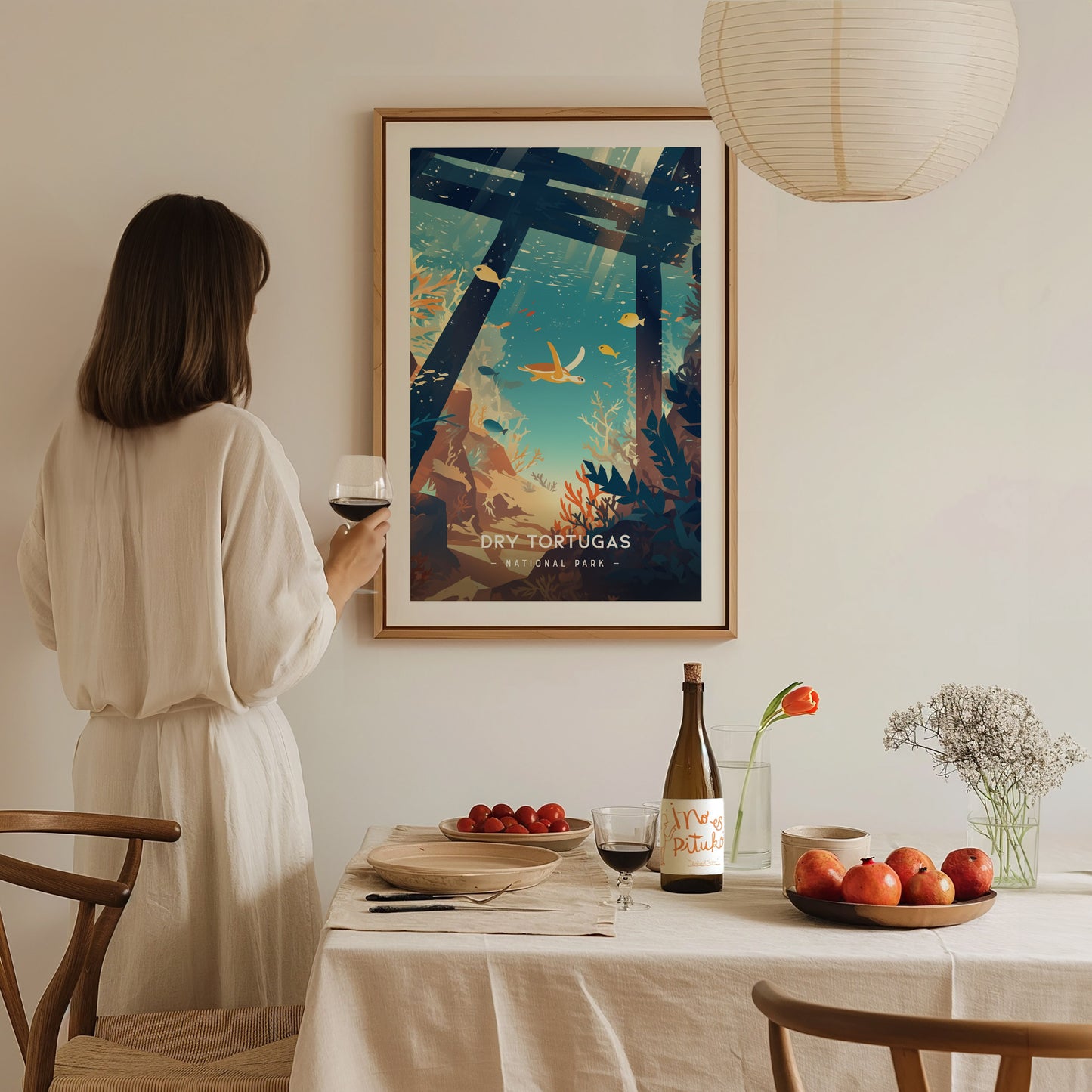 A person in a white dress holds a glass of wine while admiring a framed Dry Tortugas National Park poster. A table with fruit, wine, and flowers is in the.