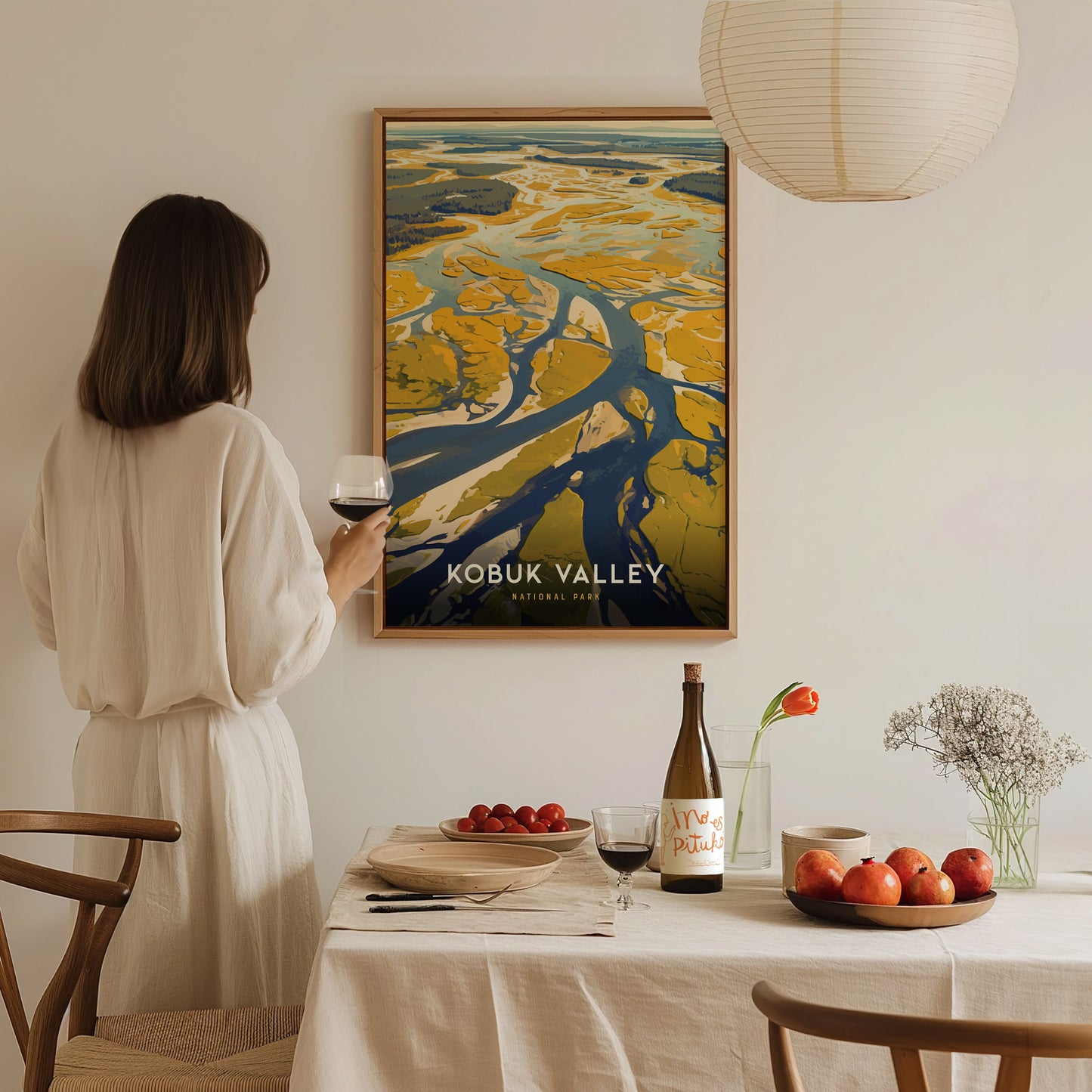 A person in a white dress holds a wine glass, admiring a Kobuk Valley National Park poster. A table with fruits, flowers, and a wine bottle is in the foreground.