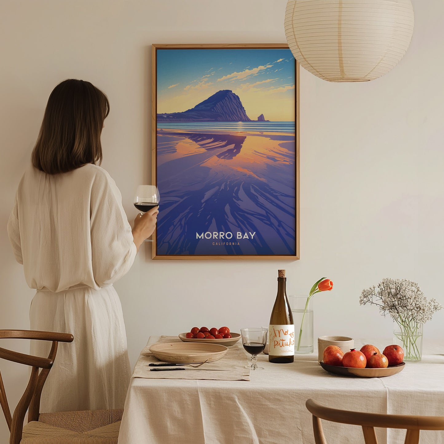 A woman in a light dress holds a glass of wine while admiring a Morro Bay poster. A table with fruit, wine, and flowers is in the foreground.