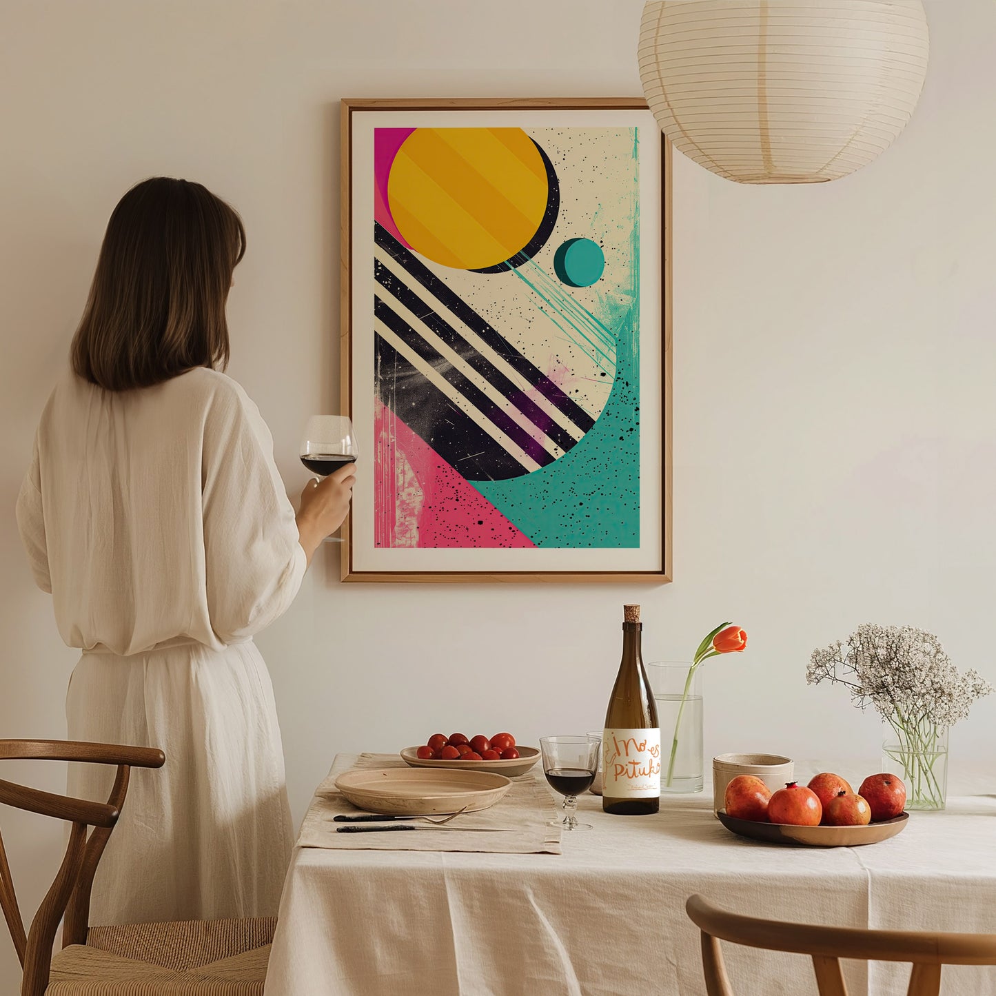A woman in a white dress holds a glass of wine, admiring a colorful 1980s abstract shapes poster. The table is set with fruit, wine, and flowers.