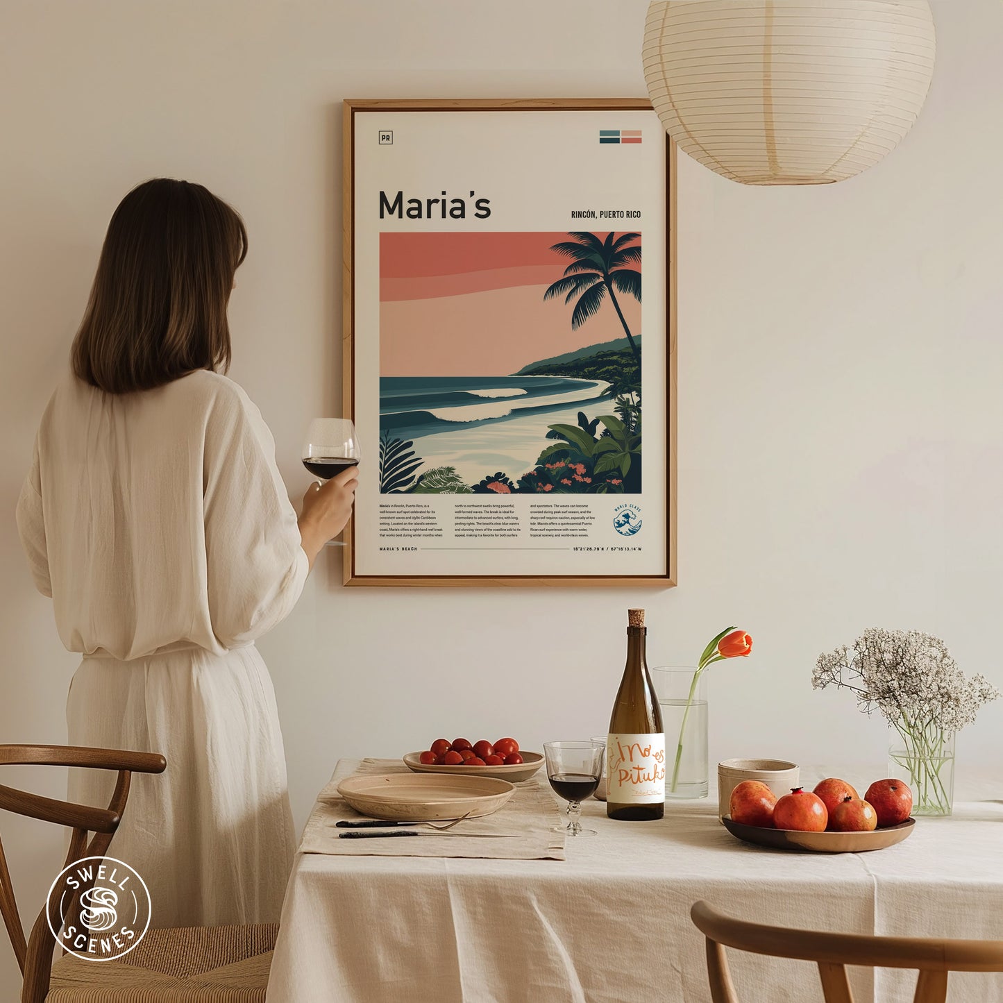 A woman in a white dress holds a glass of wine while admiring a framed surf poster on the wall. A table is set with fruit and a bottle.