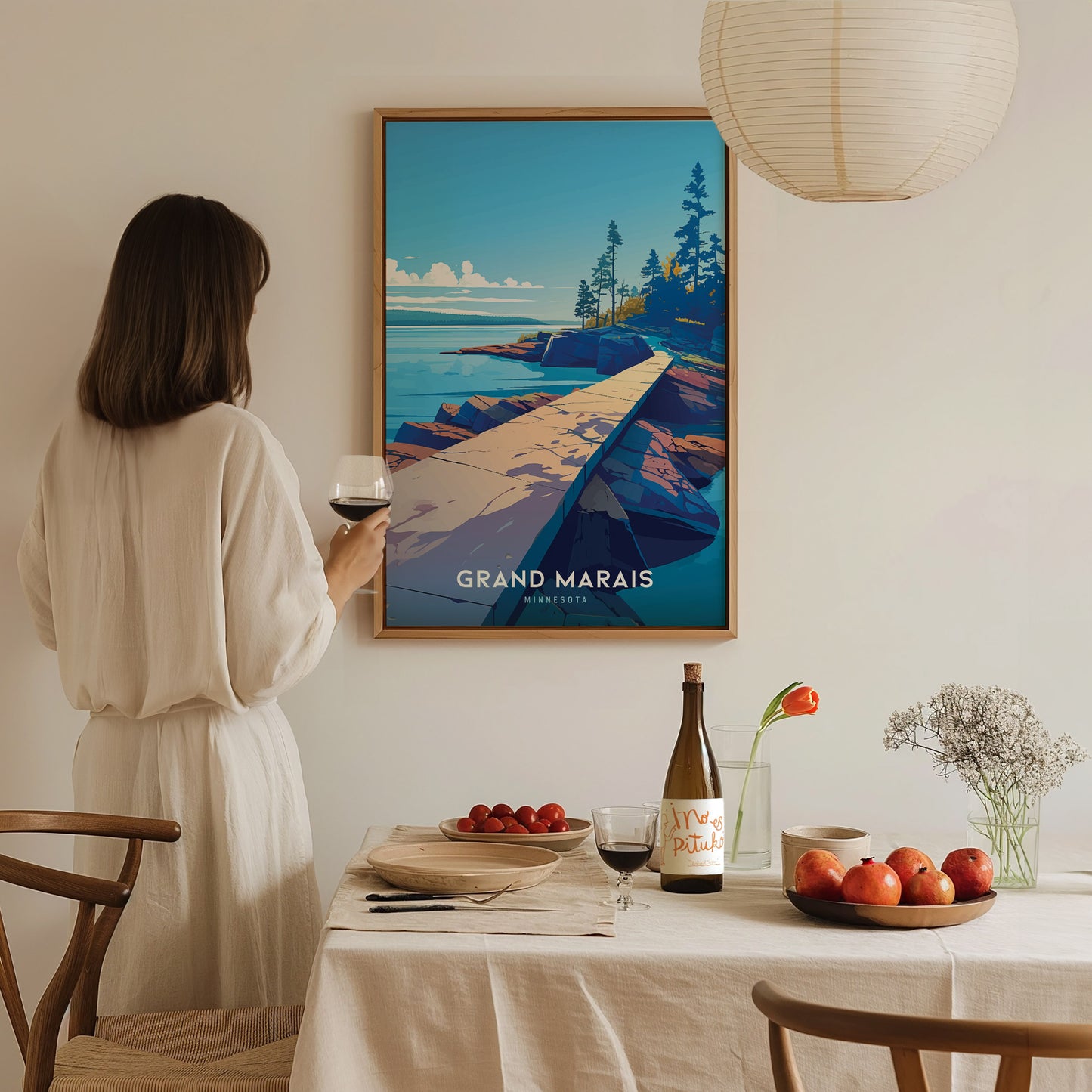 A woman in a white dress holds a glass of wine while admiring a Grand Marais, Minnesota poster. The dining table is set with fruits, flowers, and a bottle.