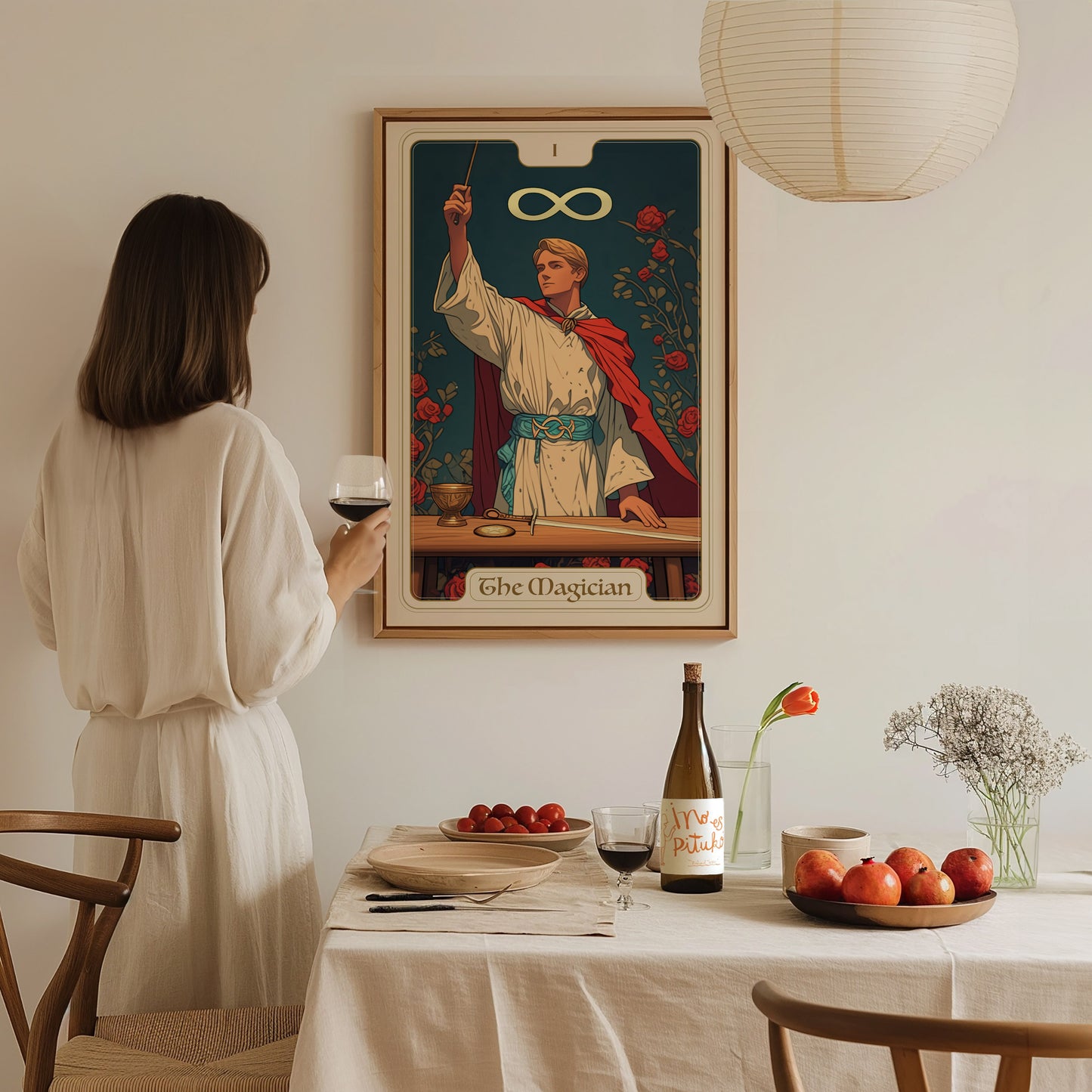 A woman in a white dress holds a wine glass and gazes at a framed Magician tarot card poster on the wall. The table is set with fruit and wine.