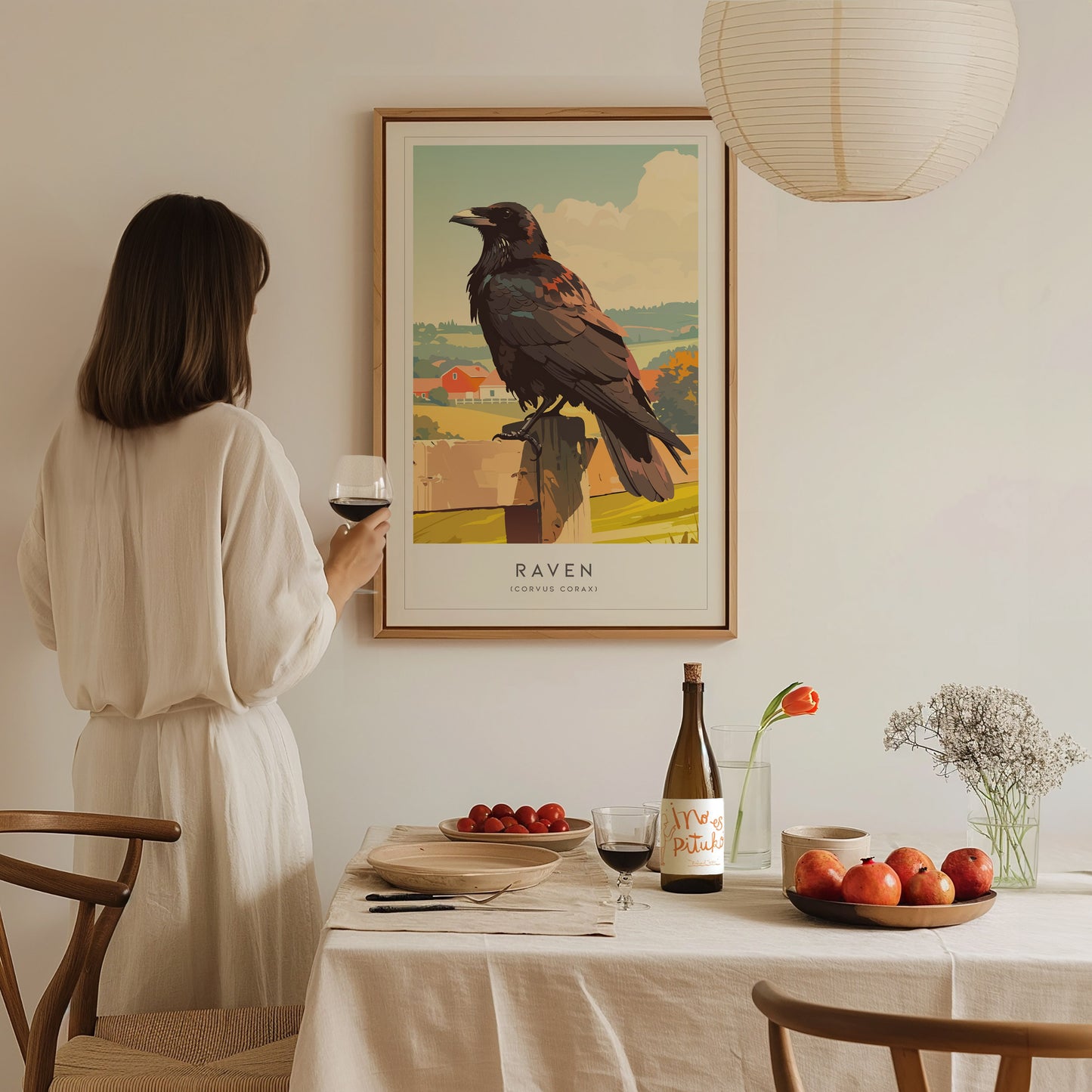 A woman in a white dress stands holding a glass of wine while gazing at a framed art print of a raven perched on a fence. The scene is set in a cozy, minimalist dining area with a table adorned with fruits and a bottle of wine.