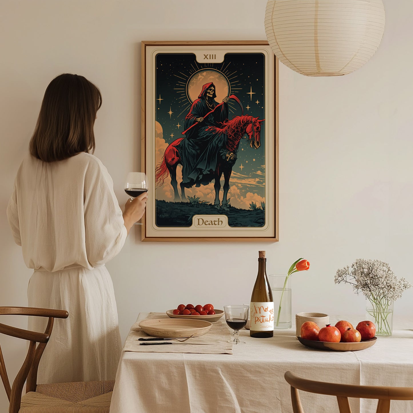 A woman in a white dress holds a wine glass, gazing at a framed Death tarot card poster on the wall. The table is set with fruits, a bottle, and.