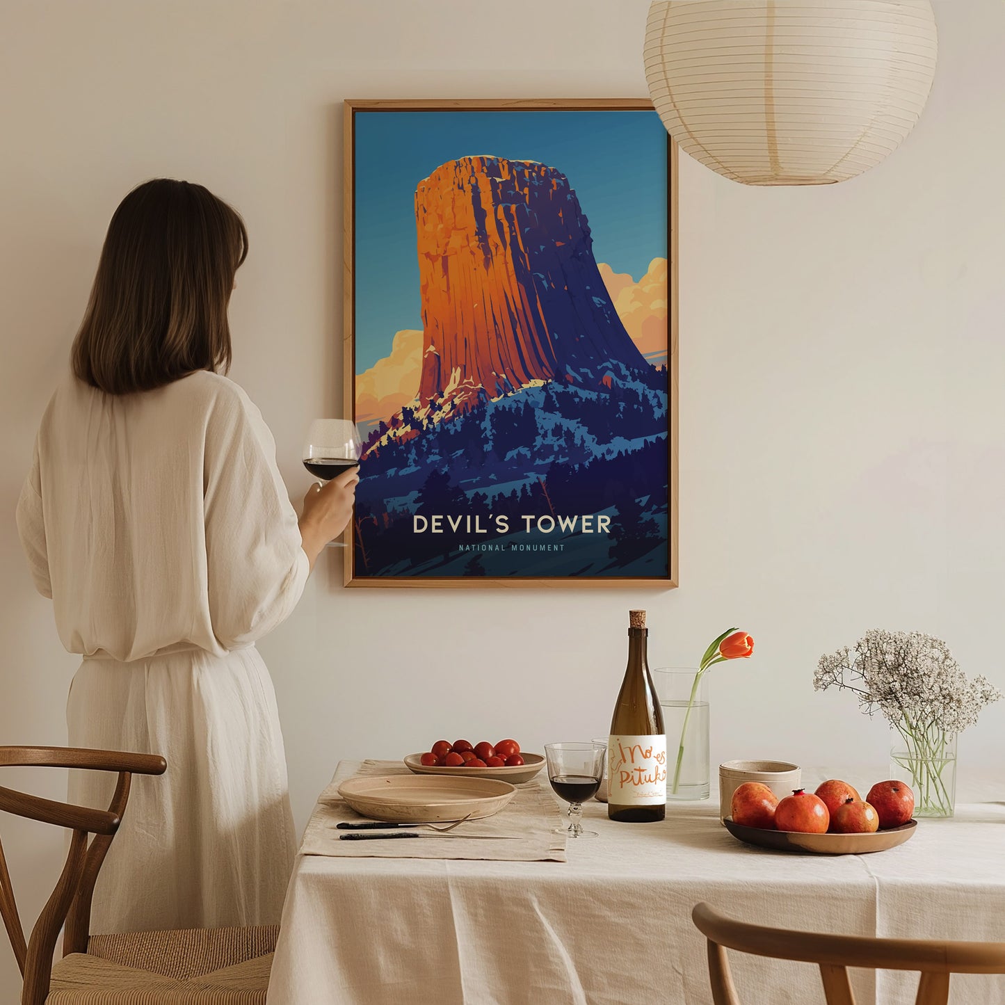 A woman in a white dress holds a glass of wine, admiring a colorful Devils Tower poster. The room features a table with fruit, flowers, and a wine bottle.