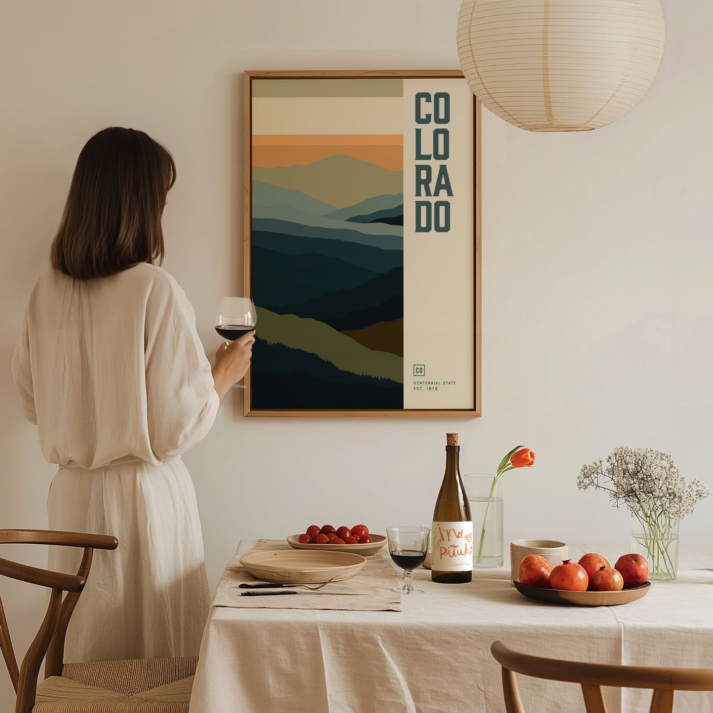 A woman in a white dress admires a framed Colorado minimalist poster featuring abstract mountain ranges. A table with wine, fruit, and flowers is set in the foreground.