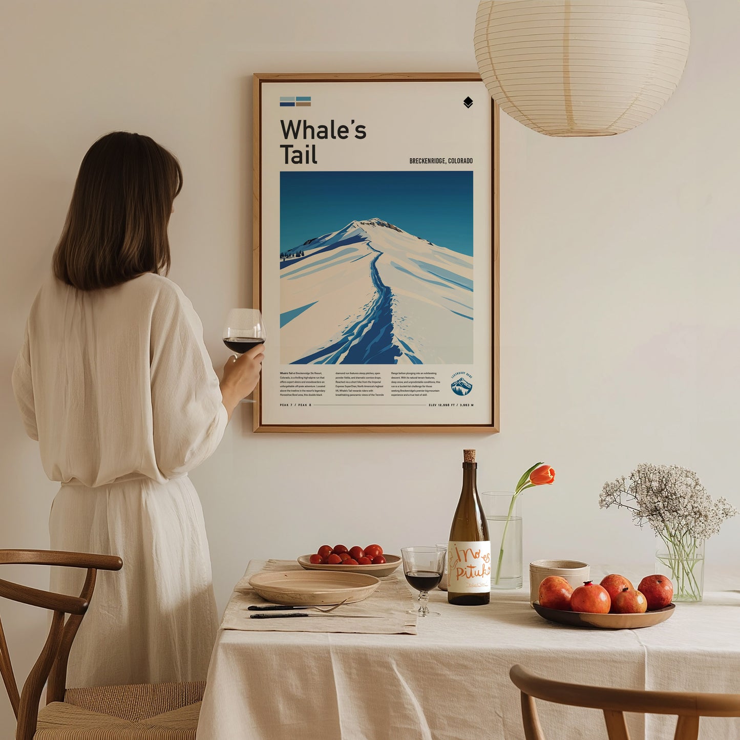 A woman in a white dress admires a framed Whales Tail poster on the wall. A dining table below features wine, apples, and a vase with flowers.
