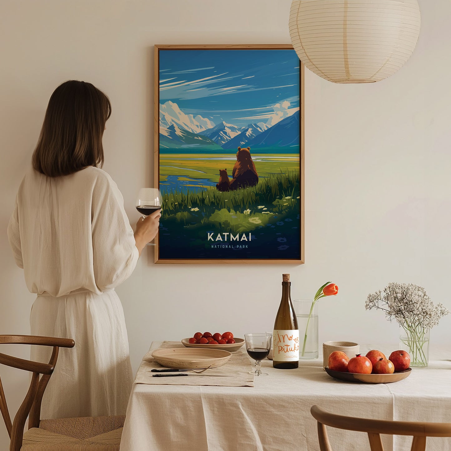 A woman in a white dress admires a Katmai National Park poster featuring bears and mountains. A dining table is set with wine, fruits, and flowers in the foreground.