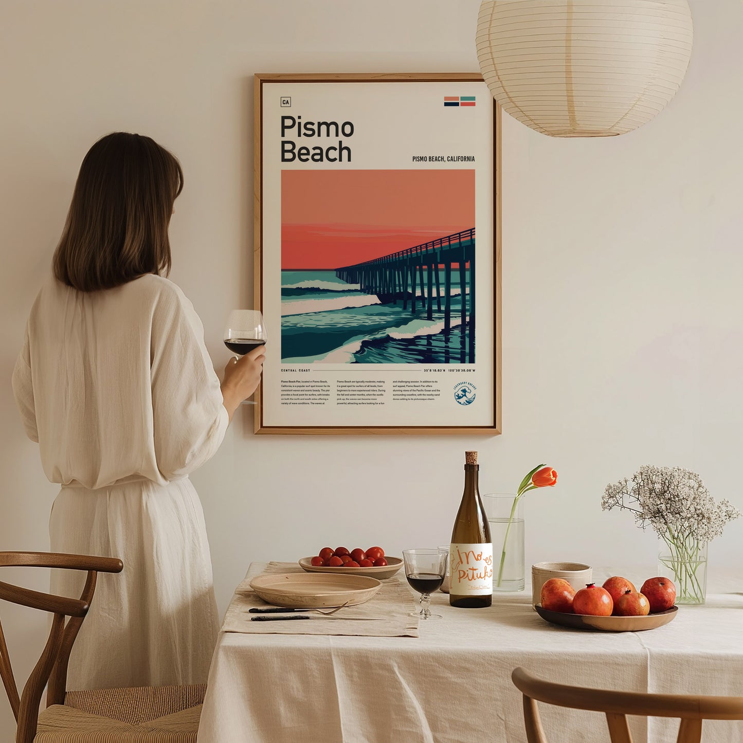 A woman in a white dress admires a framed Pismo Beach poster on the wall. A dining table with a wine bottle, apples, and flowers is in the foreground.