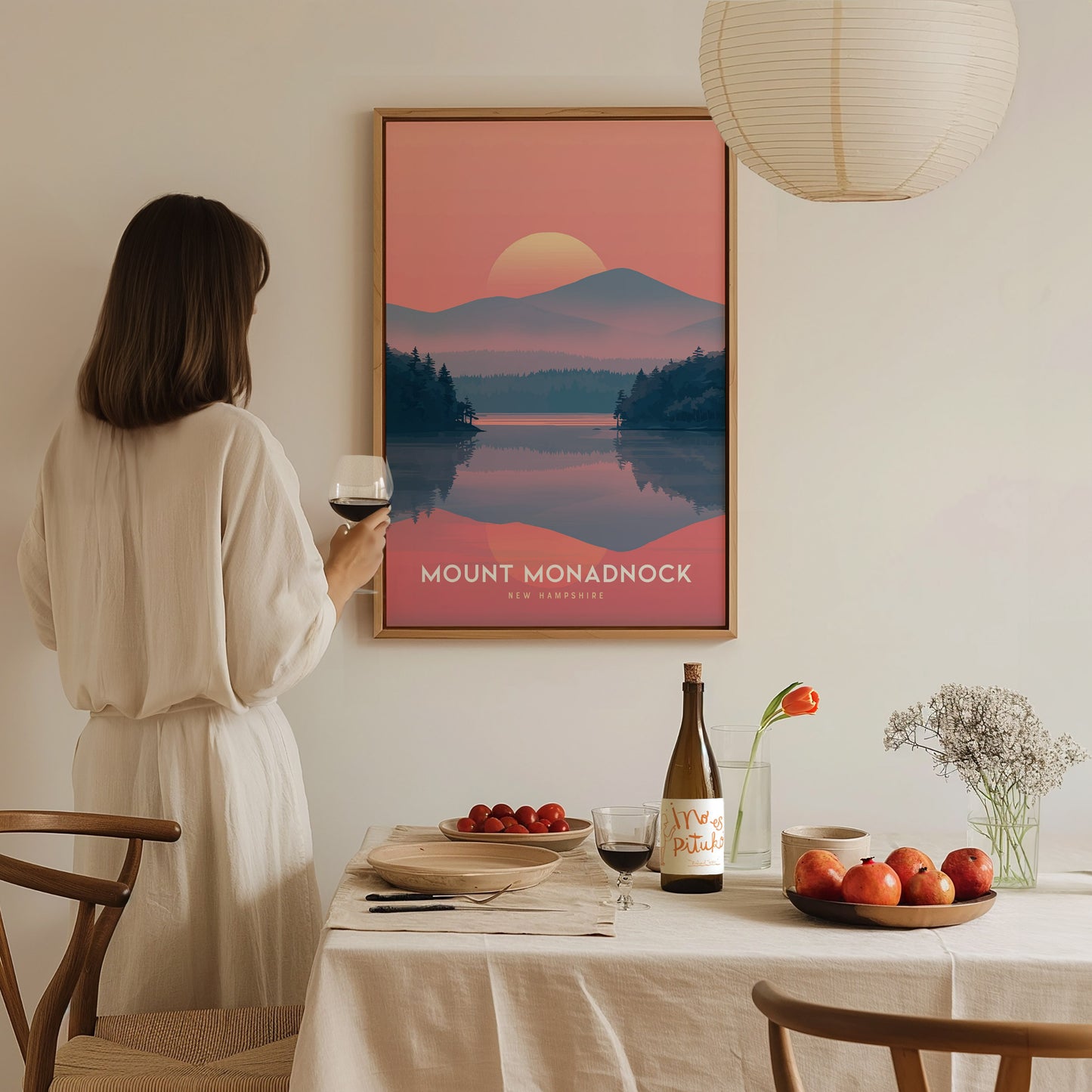 A woman in a white dress holds a glass of wine, admiring a Mount Monadnock poster on the wall. The table is set with fruit, a wine bottle, and flowers.