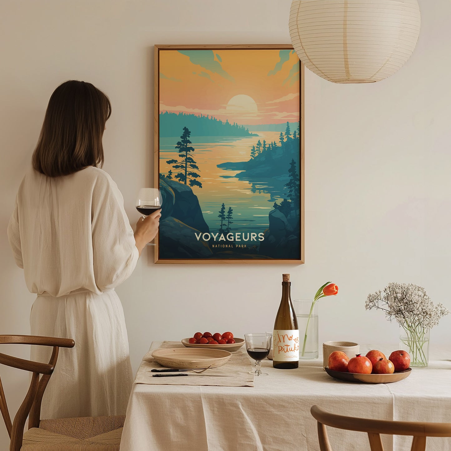 A woman in a white dress holds a wine glass while admiring a Voyageurs National Park poster. The table is set with fruit, a bottle, and a vase with flowers.