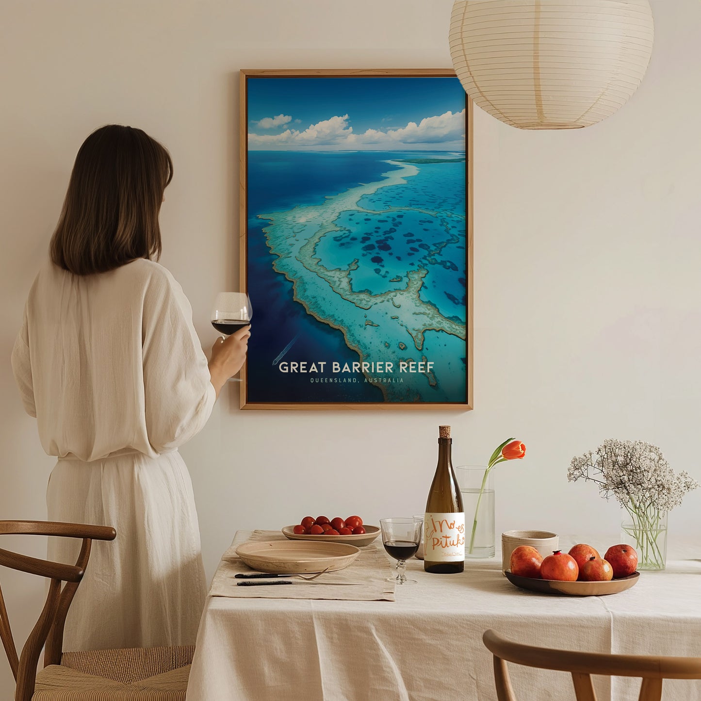 A woman in a white dress holds a wine glass, admiring a framed Great Barrier Reef poster on a wall above a set dining table with fruits and flowers.
