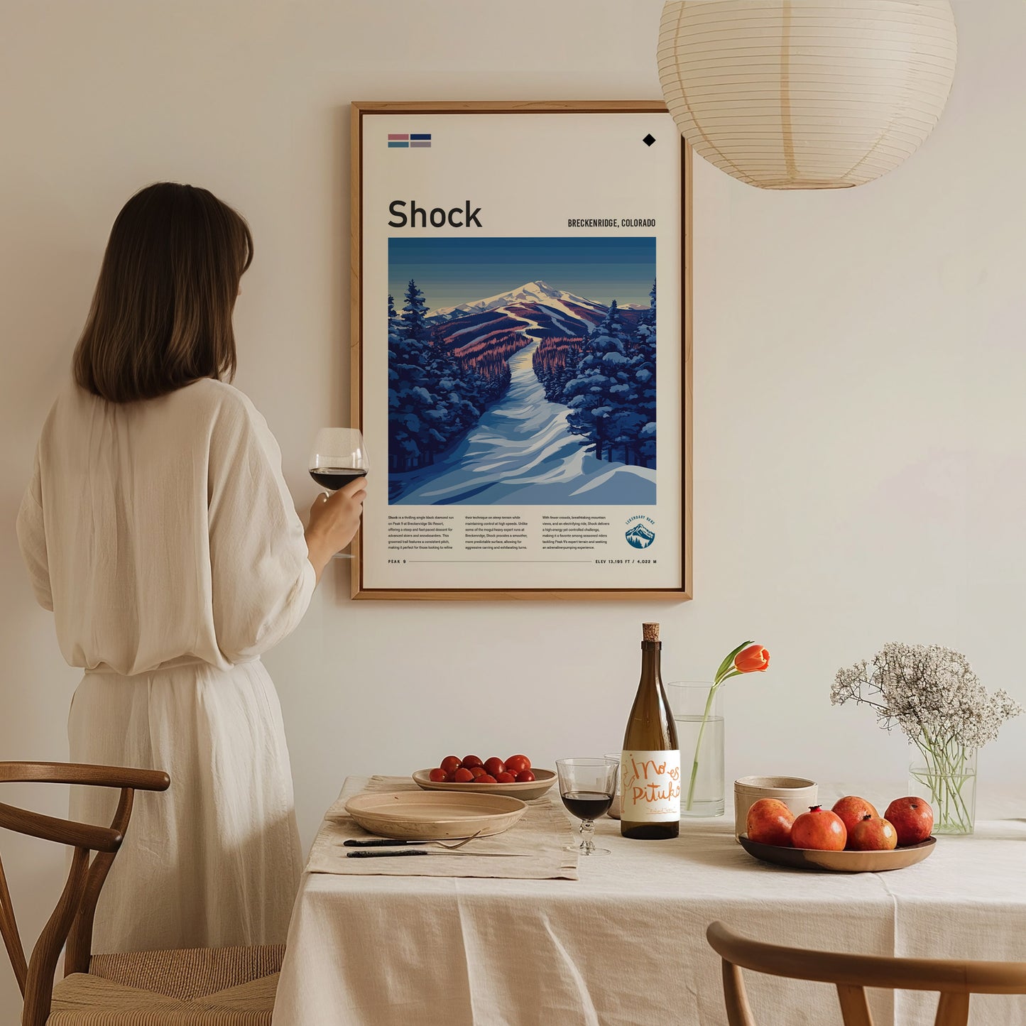 A woman in a white dress holds a glass of wine while admiring a framed ski run poster on the wall. A dining table with fruit and wine is in.
