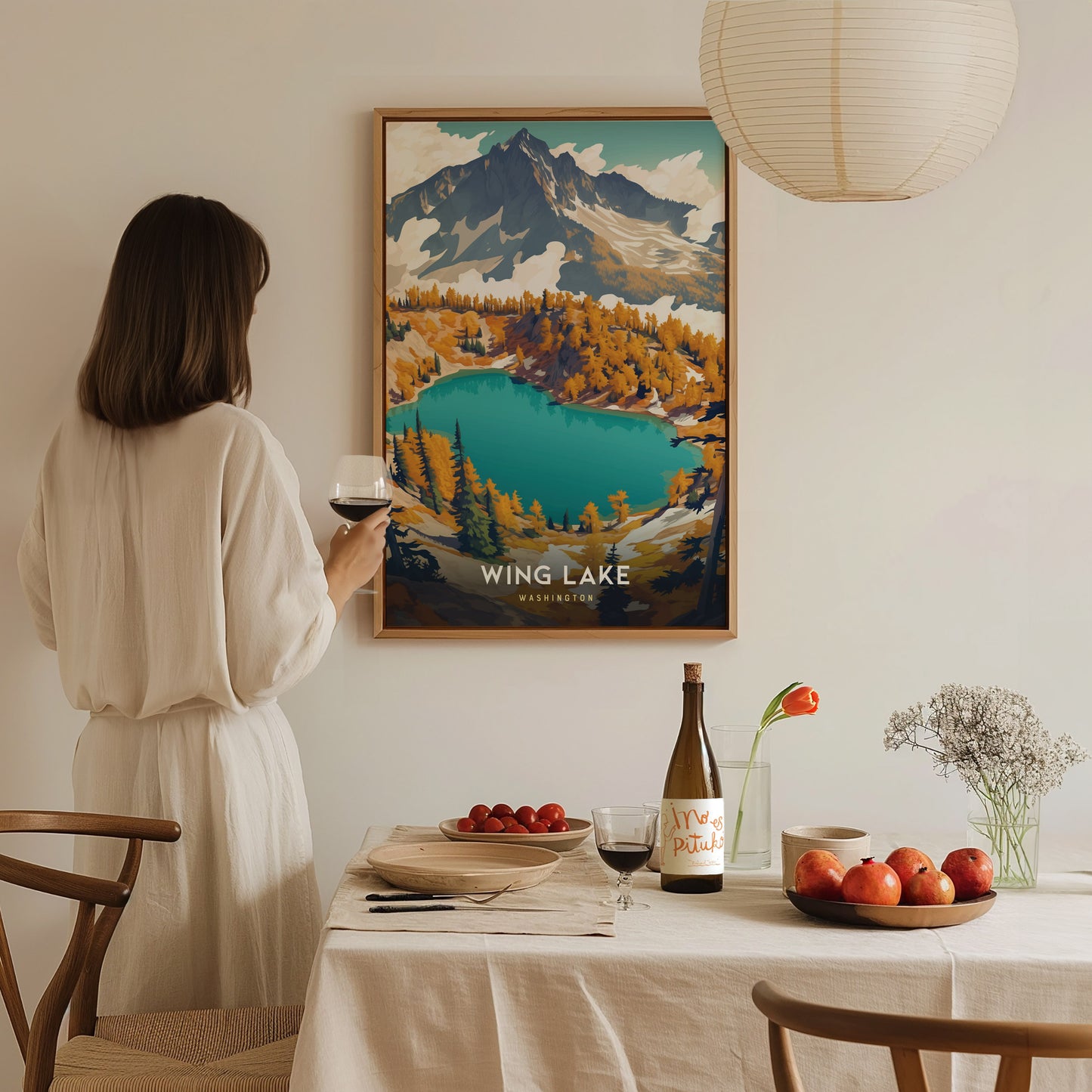 A woman in a white dress holds a glass of wine, admiring a colorful Wing Lake Washington poster on the wall. The table features a bottle, fruit, and flowers.
