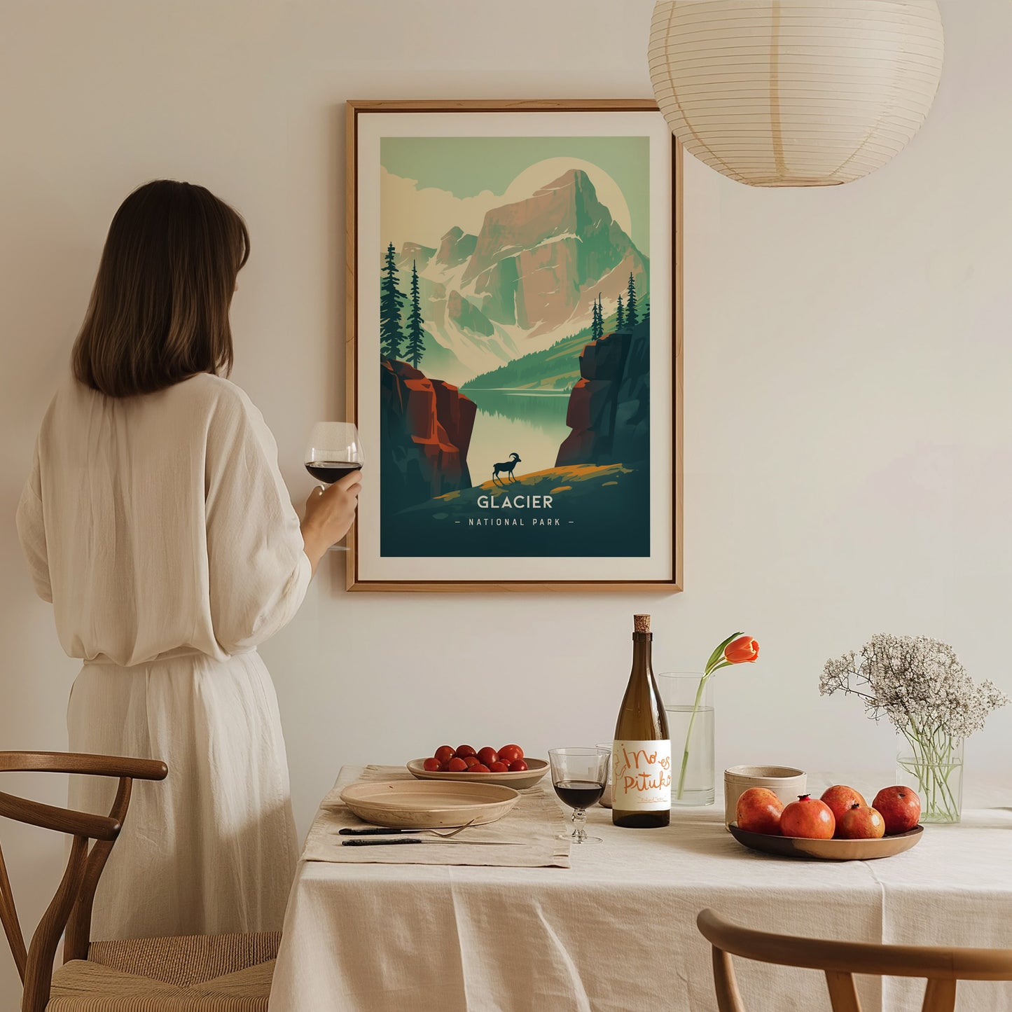 A woman in a white dress holds a glass of wine, admiring a Glacier National Park poster. A dining table with fruit, wine, and flowers is in the foreground.