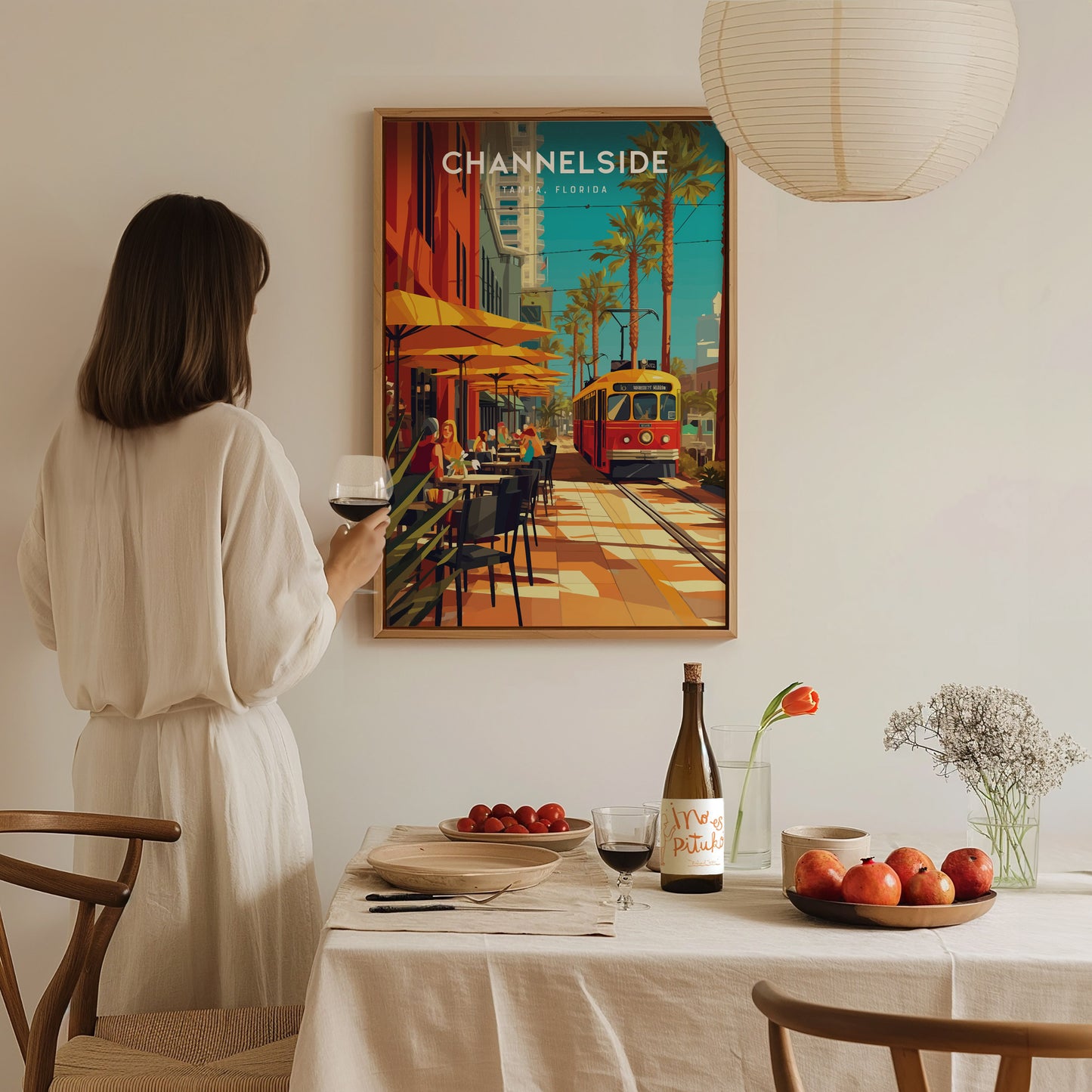 A woman in a white dress admires a vibrant Channelside Tampa poster featuring a red trolley. A table with drinks, fruit, and flowers is set in the foreground.