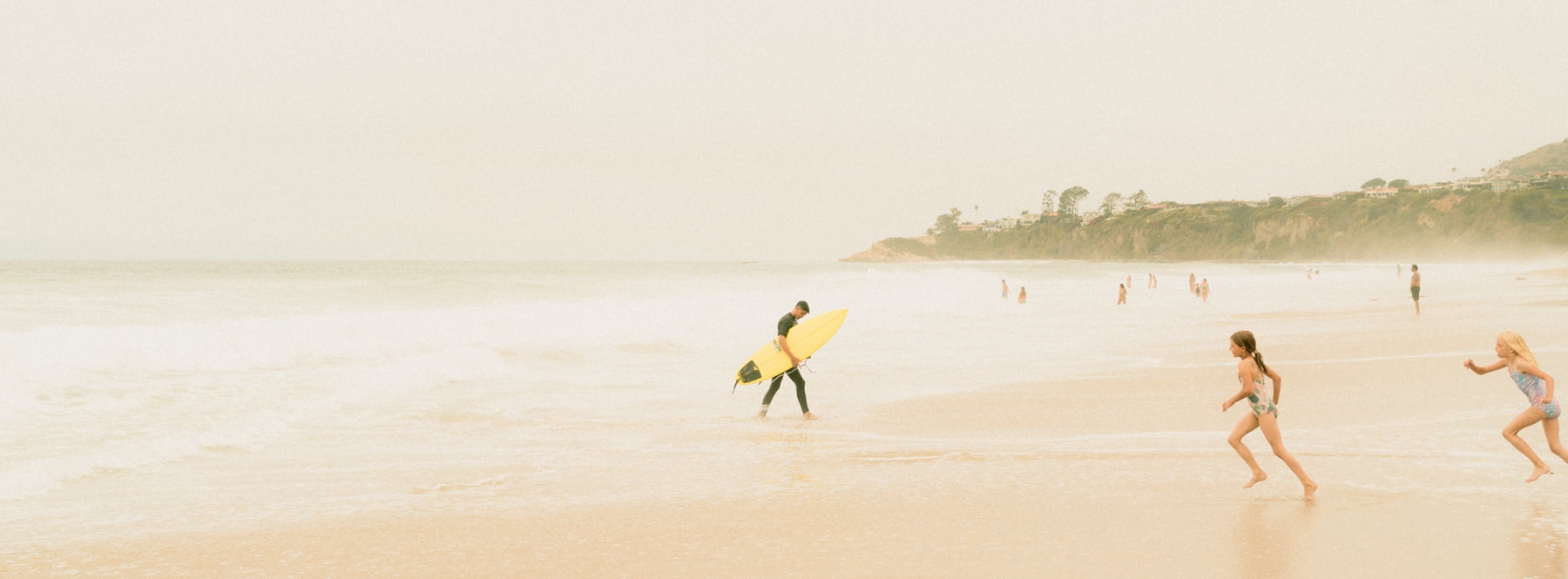 A surfer walks on the beach while children run.