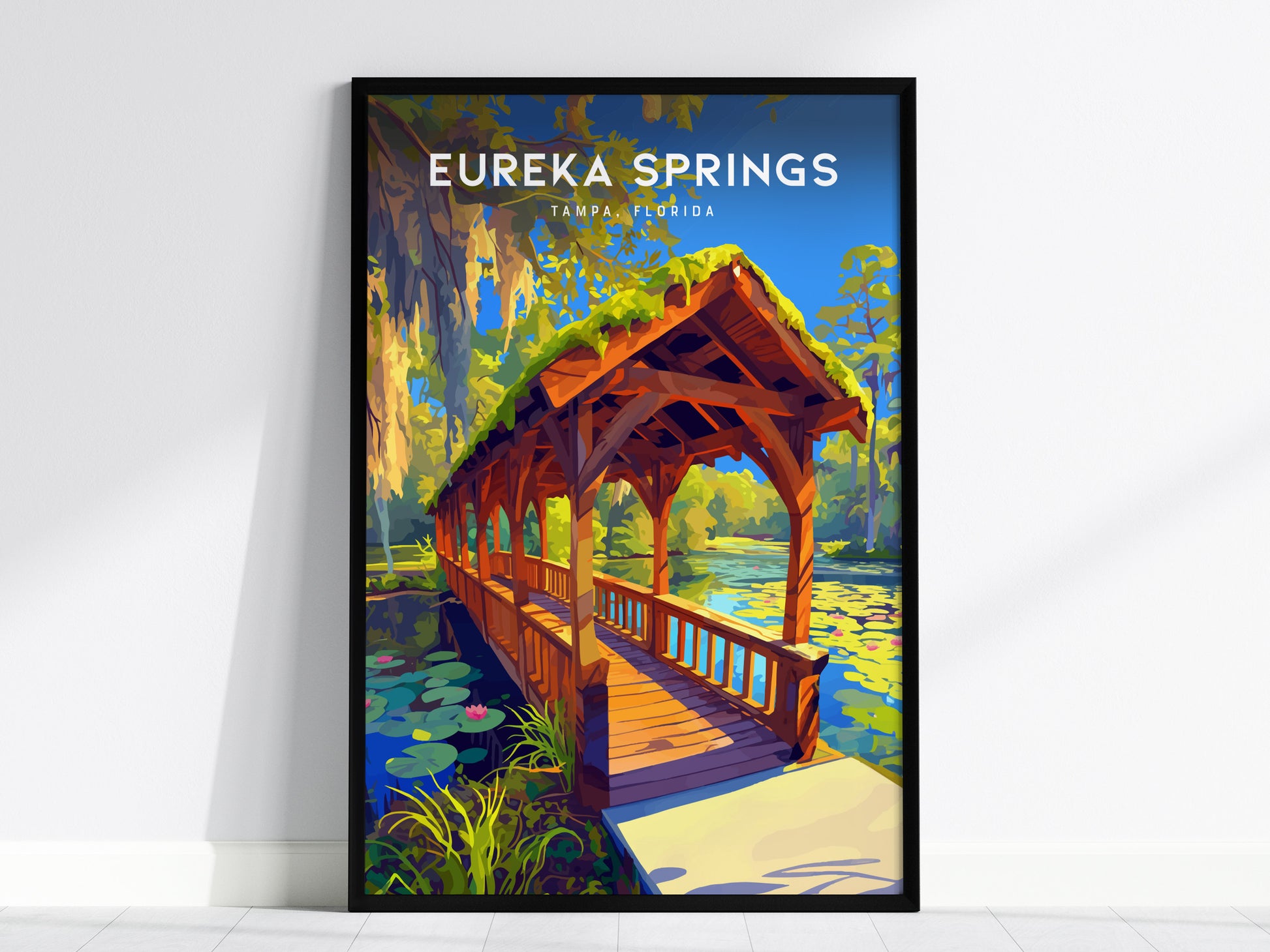 A framed poster of Eureka Springs in Tampa, Florida, featuring a covered wooden bridge over a serene stream, surrounded by vibrant greenery and water lilies.