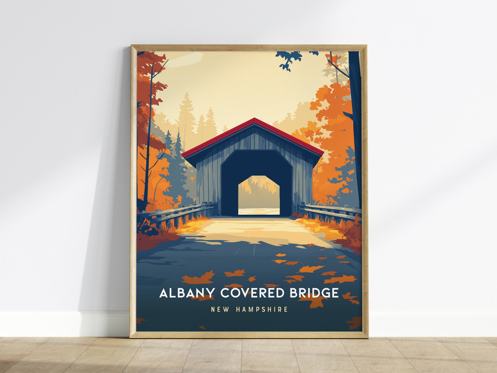 A framed poster of the Albany Covered Bridge in New Hampshire is displayed against a white wall. The artwork features the bridge set amidst vibrant autumn foliage with rich oranges and yellows, capturing the essence of a serene fall.