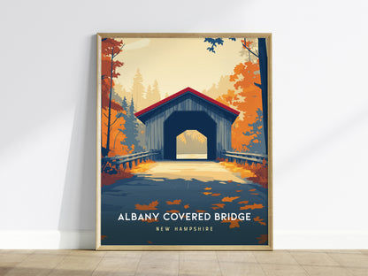 A framed poster of the Albany Covered Bridge in New Hampshire is displayed against a white wall. The artwork features the bridge set amidst vibrant autumn foliage with rich oranges and yellows, capturing the essence of a serene fall.