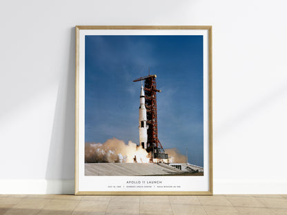Framed poster of Apollo 11 launch with a rocket on a launch pad against a blue sky.