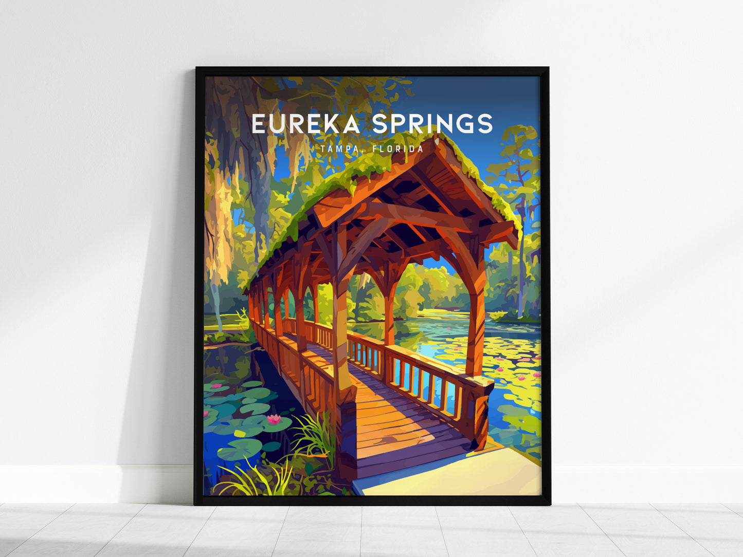 A framed poster of Eureka Springs, Tampa, depicts a covered wooden bridge over a tranquil stream surrounded by lush greenery and water lilies under a clear blue sky.
